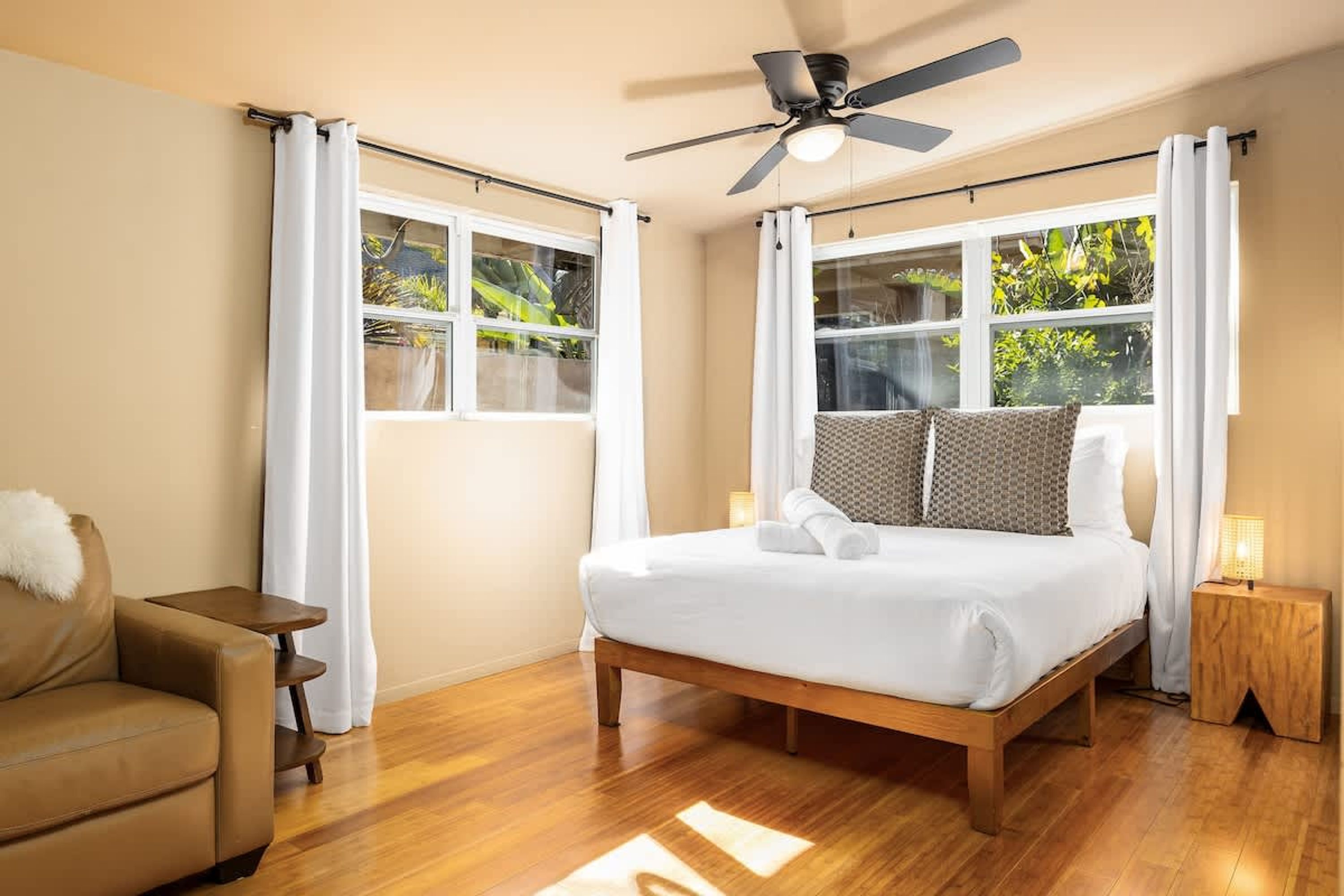 Une chambre à coucher comprenant un lit, un ventilateur de plafond, une table de chevet, une chaise et de grandes fenêtres avec des rideaux.