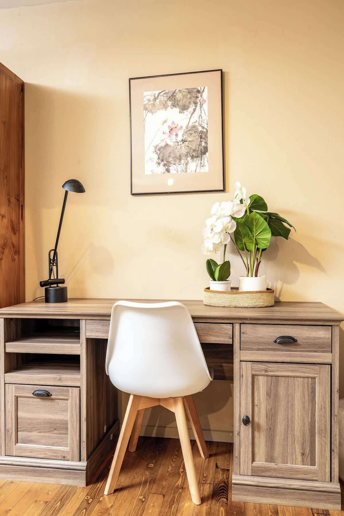 Een houten bureau met een witte stoel, een lijst met kunstwerk, een vloerlamp en een potplant met witte bloemen.