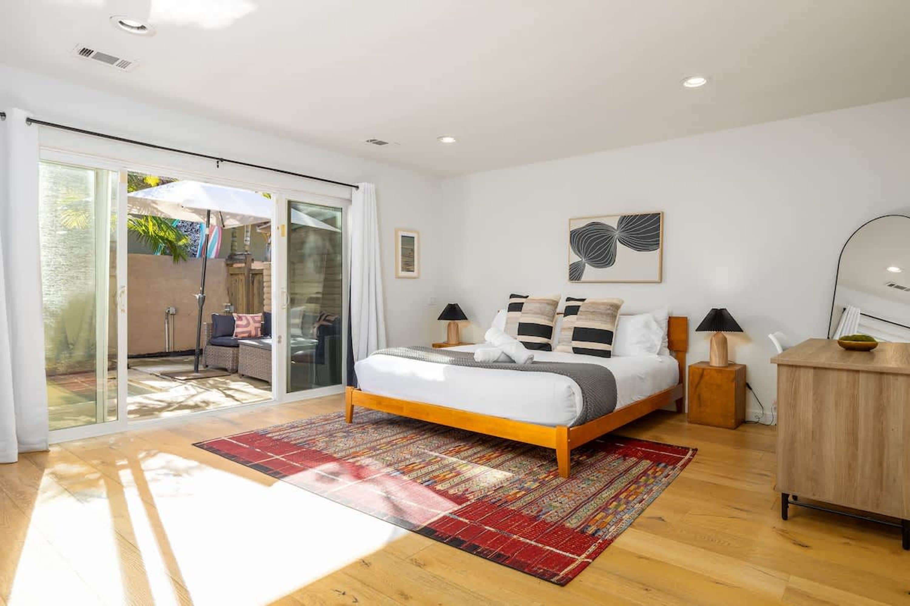 A modern bedroom with a large bed, a red patterned rug, a wooden cabinet, and a sliding glass door leading to an outdoor area.