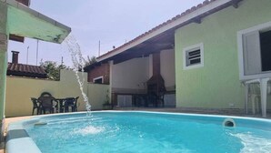 Una piscina con chorros de agua, un patio con sillas y un edificio con tejado de baldosas.