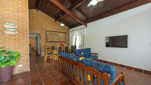 Una habitación con pared de ladrillo, vigas de madera en el techo, zona de comedor con sillas y mesa, un conjunto de sofás, televisor montado en la pared y un cuadro grande.