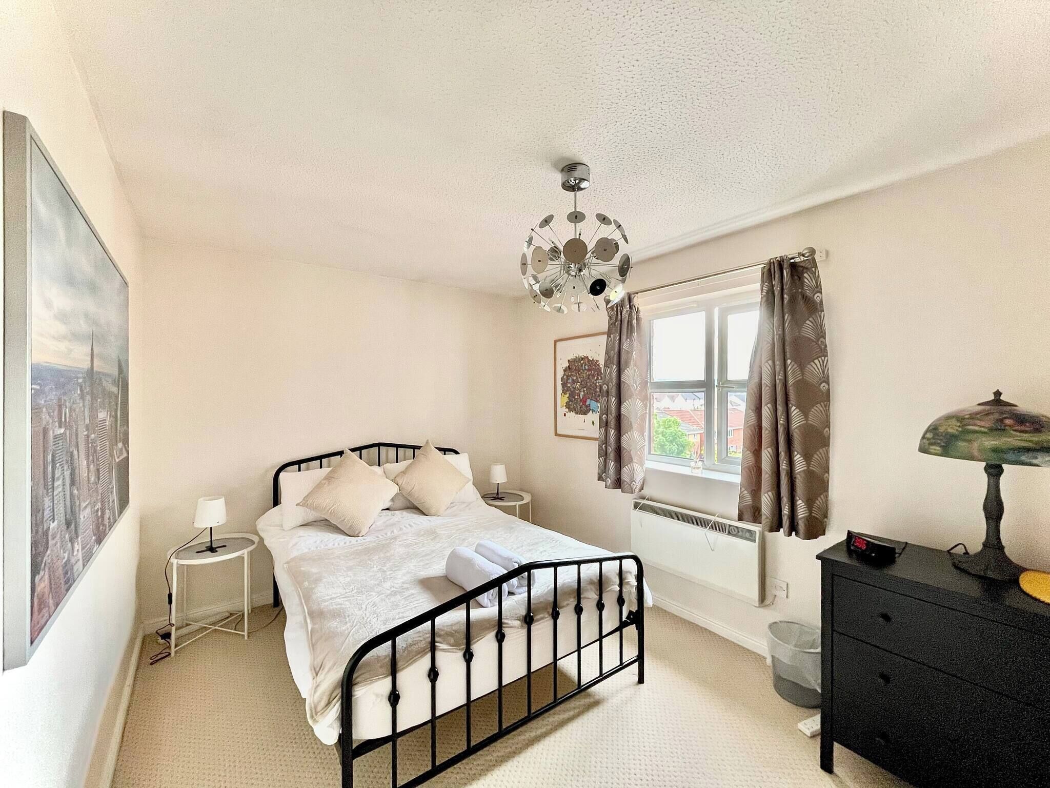 Een slaapkamer met een enkel bed, een nachtkastje, een lamp, een radiator, een raam met gordijnen en een aan de muur hangend schilderij.