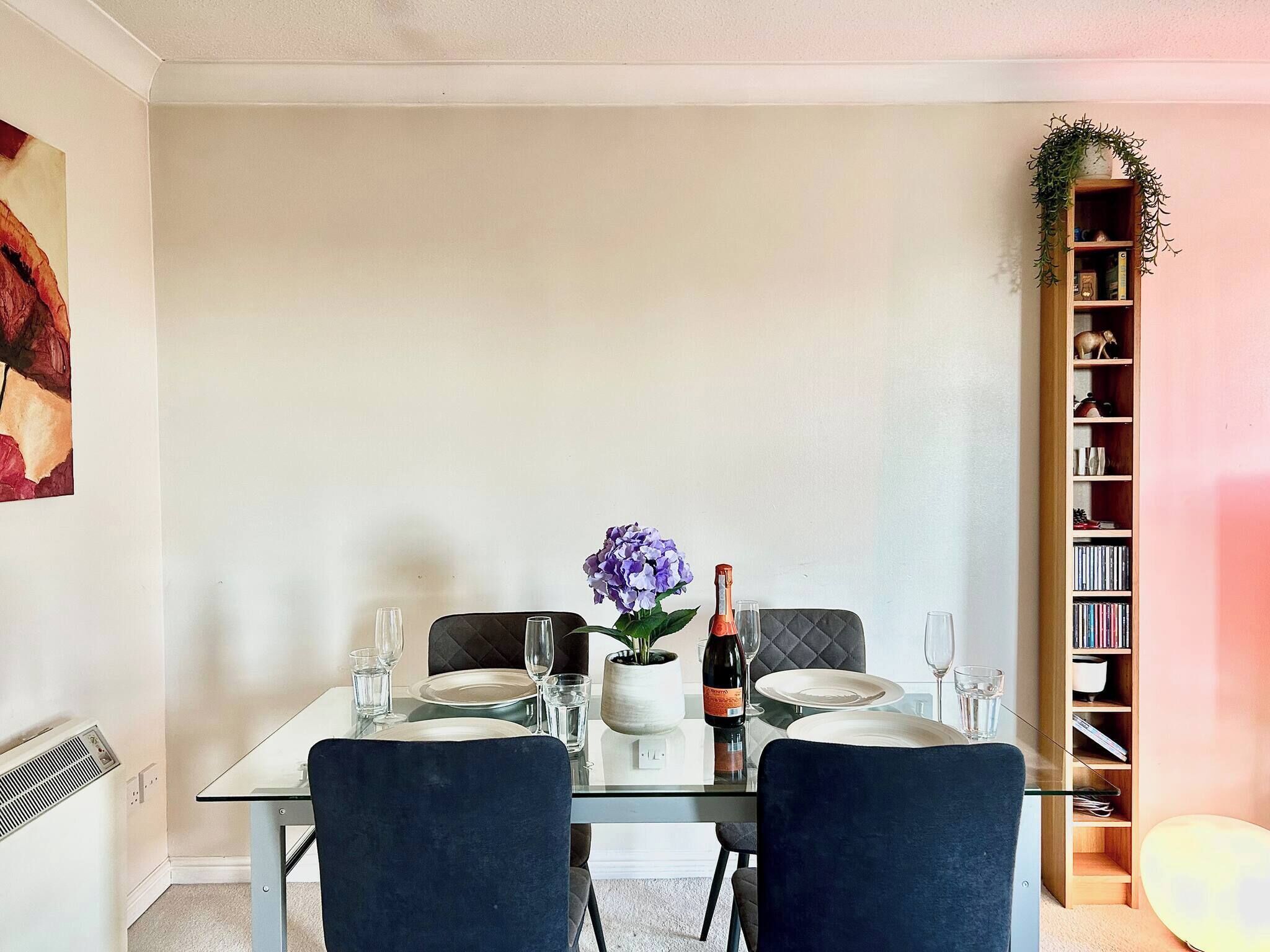 Un comedor con una mesa de vidrio dispuesta para dos personas, un florero con flores moradas y una botella de vino.