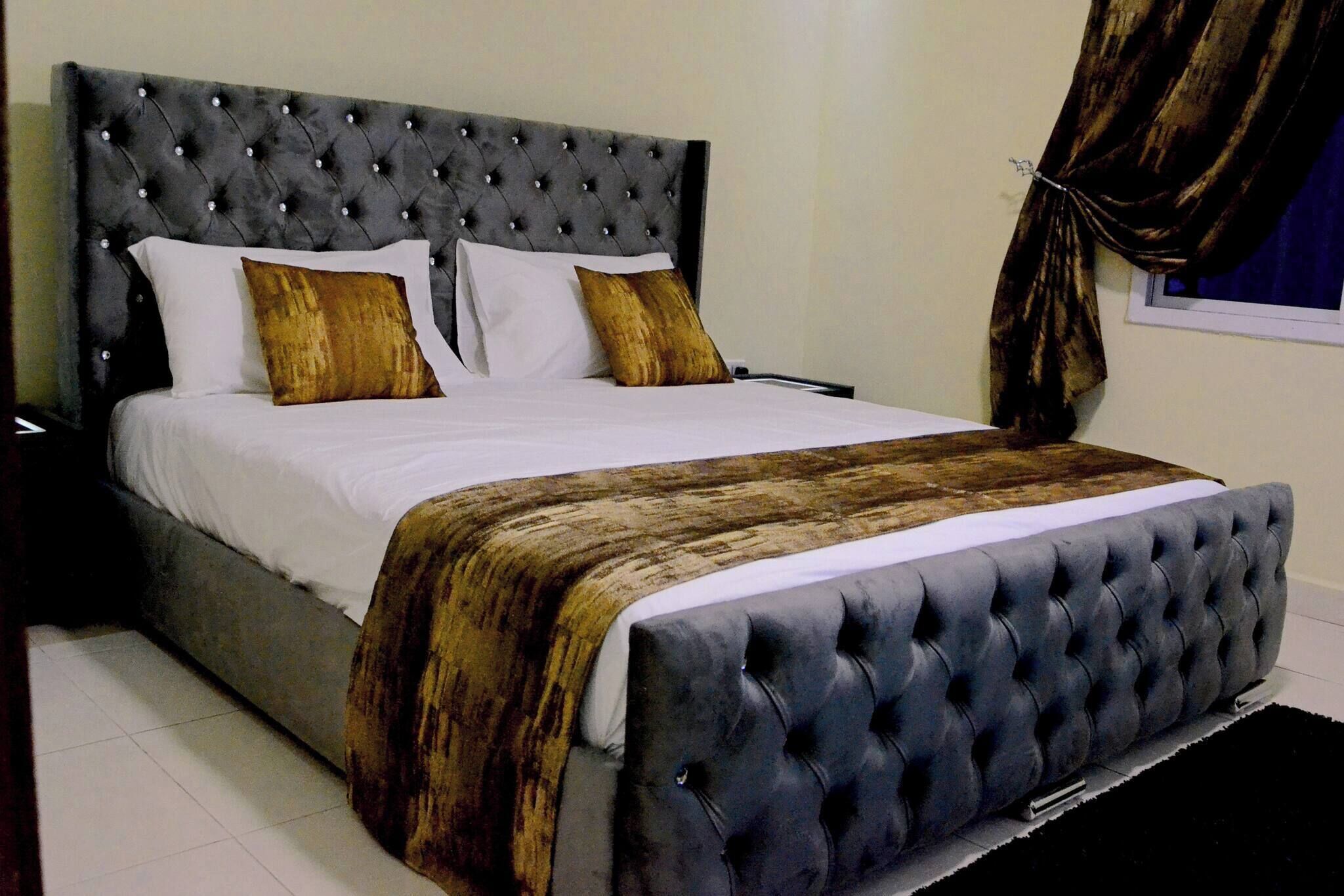 Cama com cabeceira estofada, roupa de cama branca e duas almofadas decorativas.