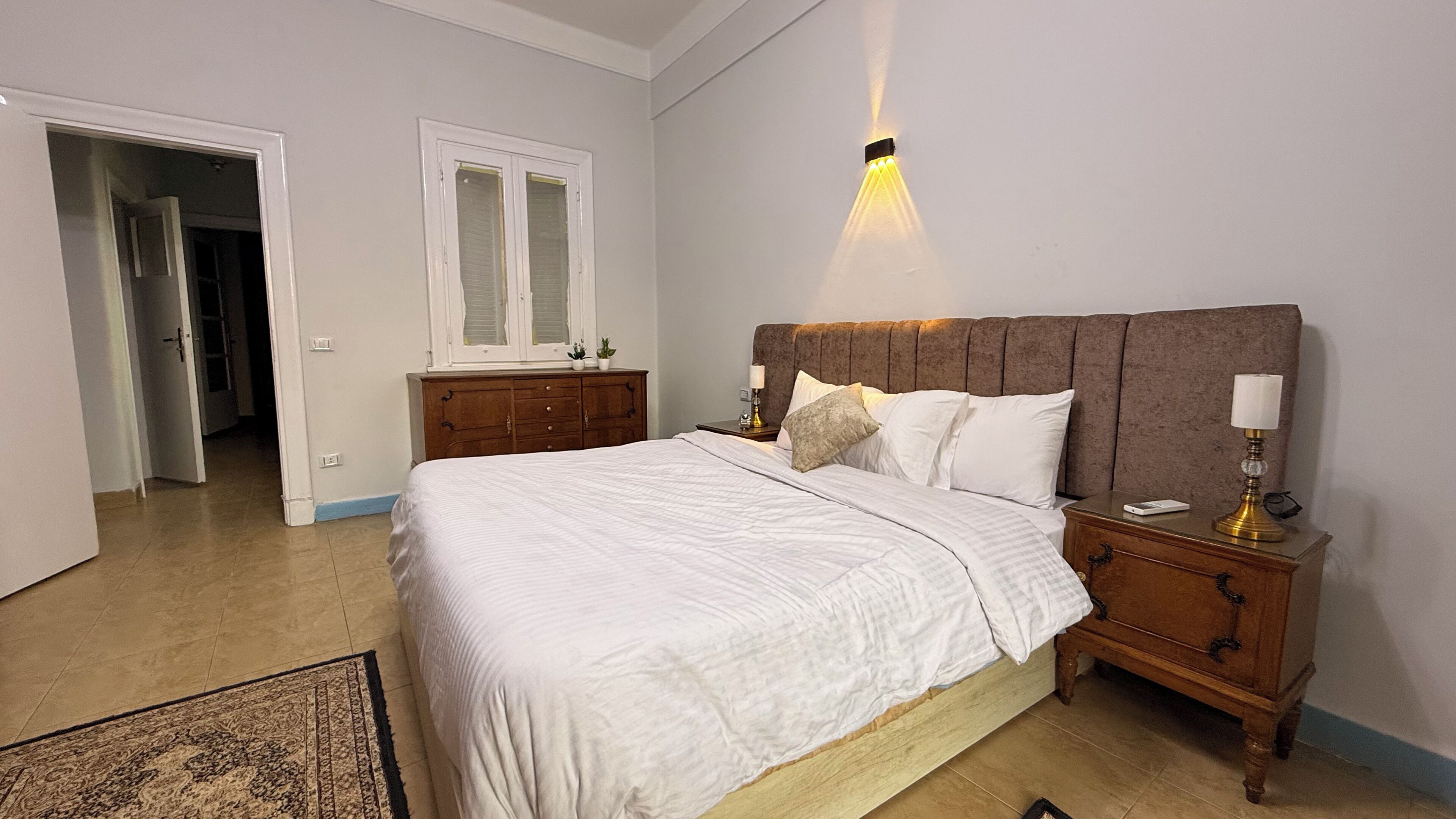 Ein Schlafzimmer mit einem großen Bett, einem Nachttisch, einer Kommode und einem Fenster mit Jalousien.