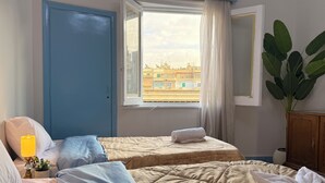 A hotel room with two beds, a window with a view of buildings, a potted plant, and a wooden cabinet. - Palace View Royal Home (Cairo)