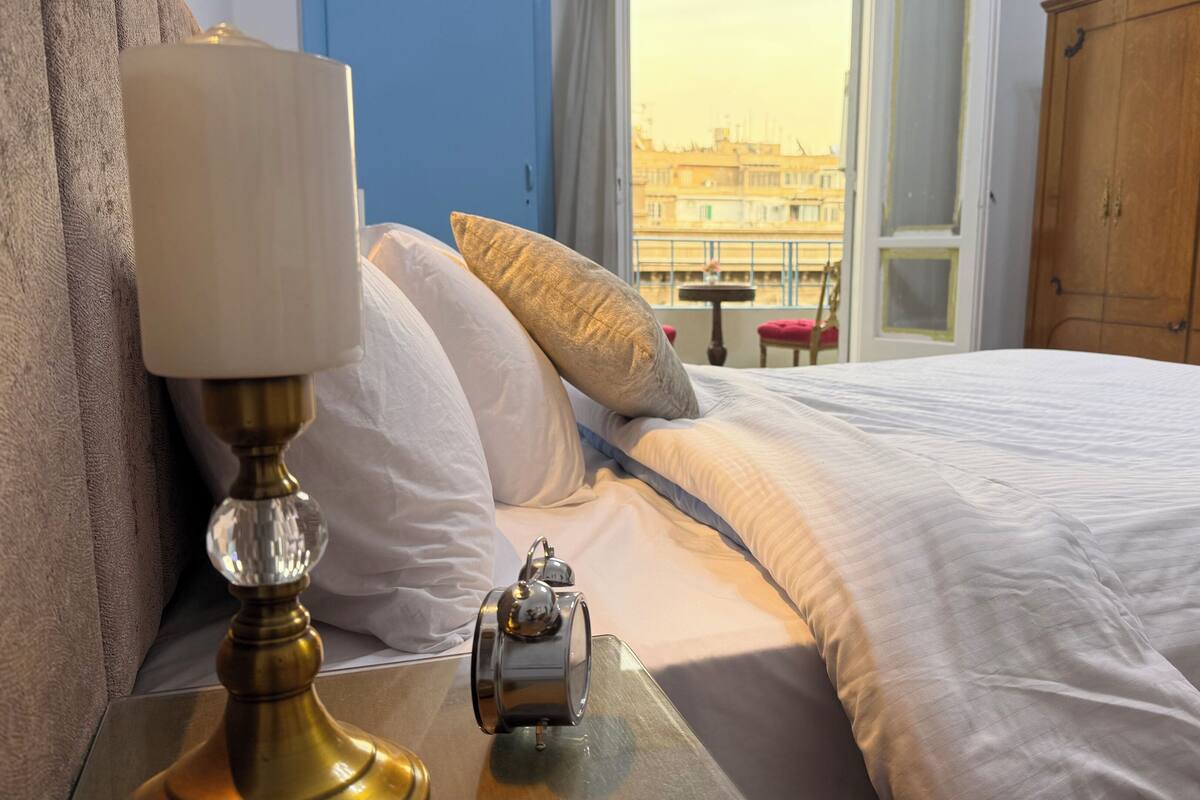 Un letto rifatto con lenzuola bianche, una lampada in ottone e un tavolino con una sveglia. Una finestra mostra una vista sulla città.