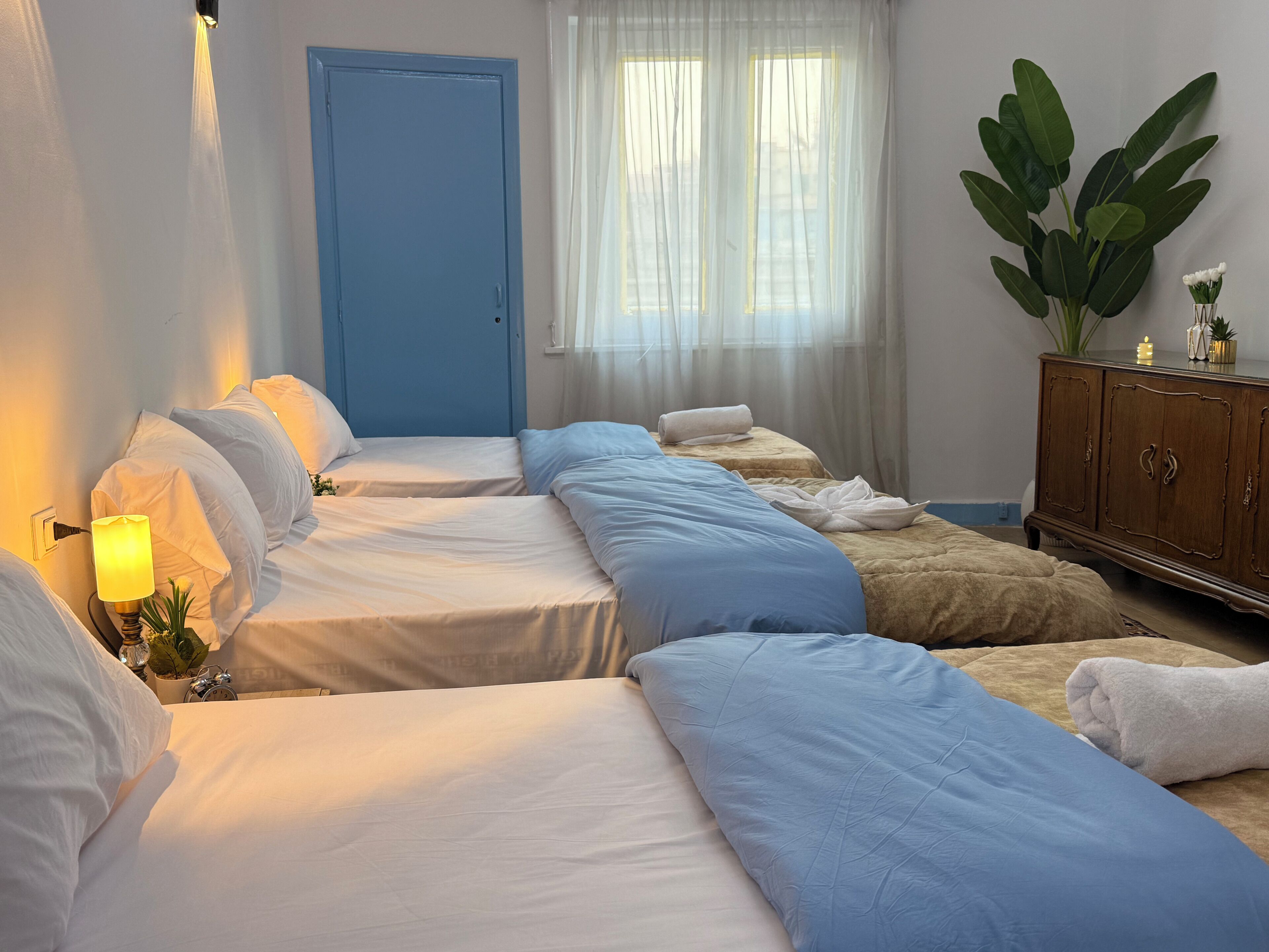 A hotel room with two single beds, a wooden cabinet, a potted plant, and a window with sheer curtains.