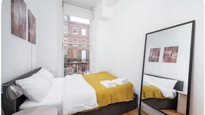 A modern hotel room with a bed, bedside table, and a large window offering a city view. - Flat 1 Modern Marylebone Apartment with Balcony (London)