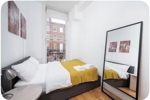 A modern hotel room with a bed, bedside table, and a large window offering a city view. - Flat 1 Modern Marylebone Apartment with Balcony (London)