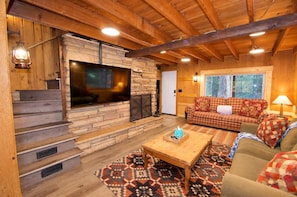A cozy living room with a stone fireplace, a large TV, a checkered sofa, and a patterned rug. - Creekside Cabin by Cottonwood Lodging (Brighton)
