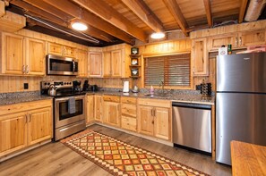 A kitchen with wooden cabinets, stainless steel appliances, and a patterned rug. - Creekside Cabin by Cottonwood Lodging (Brighton)