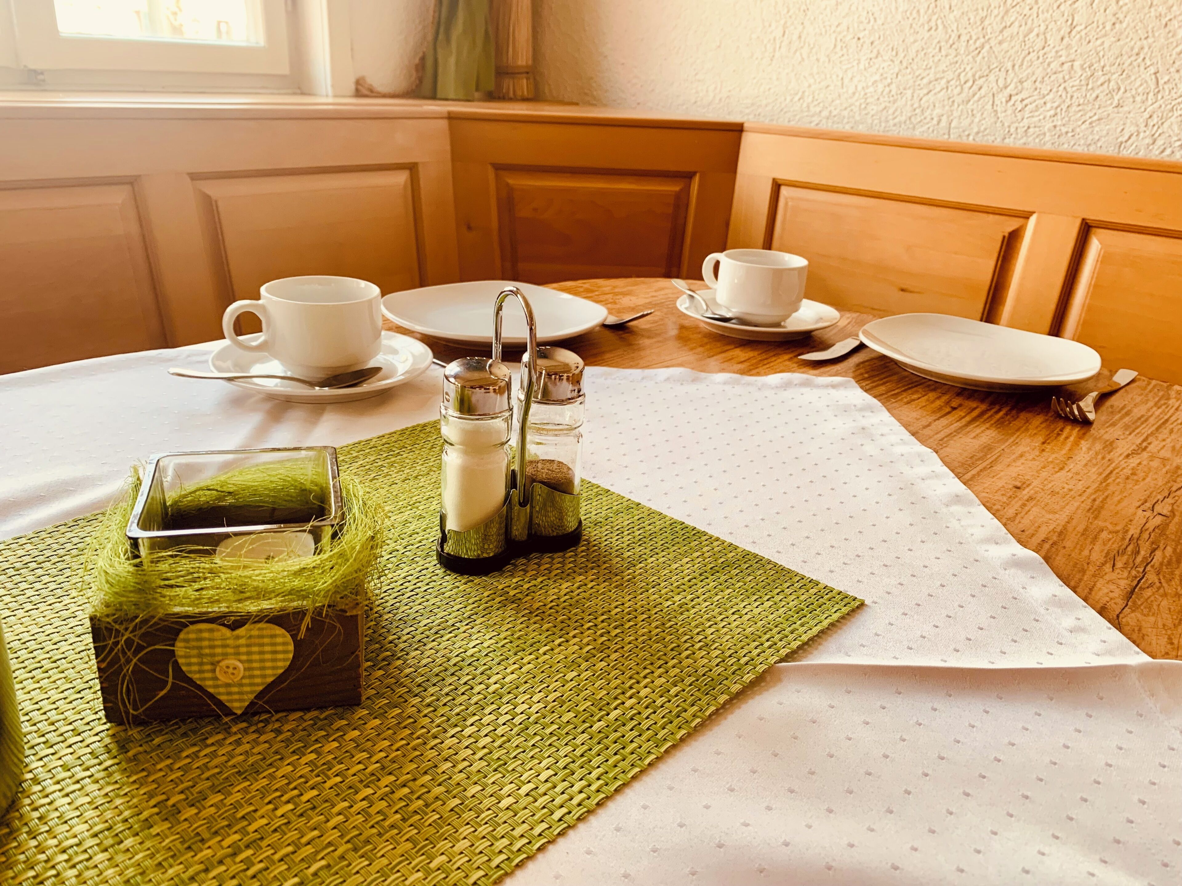 A dining table with a white tablecloth, two salt and pepper shakers, a glass container with a heart design, and a woven placemat.
