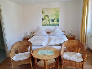 Quarto com cama, duas mesas de cabeceira, um quadro, duas cadeiras de vime e uma pequena mesa com um copo.