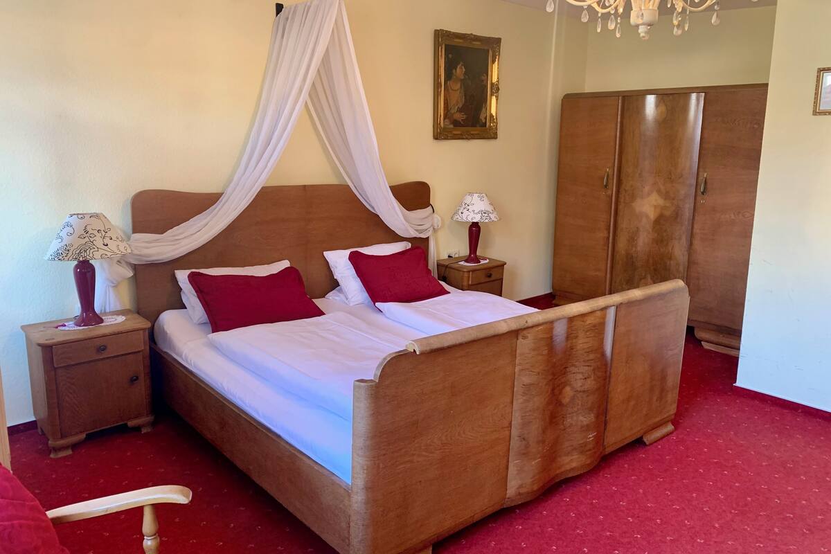 A bedroom with a wooden bed, red pillows, a nightstand with a lamp, a wardrobe, and a chandelier.