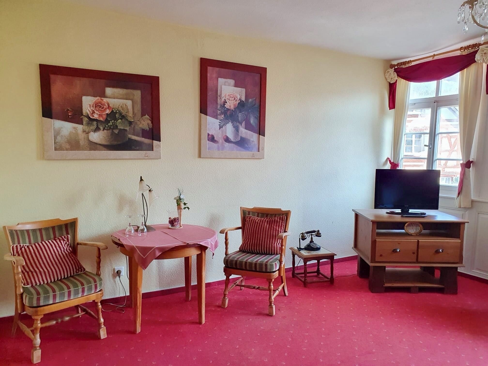 Una habitación con alfombra roja, dos sillas de madera con cojines a rayas, una mesita con mantel rojo, un televisor sobre un mueble de madera y dos cuadros enmarcados en la pared.
