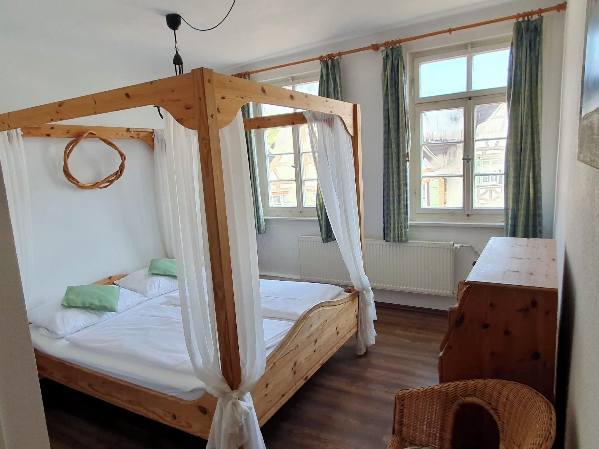 Una cama con dosel de madera con ropa de cama blanca, una silla de mimbre y una pequeña mesa de madera en una habitación con ventanas y cortinas.