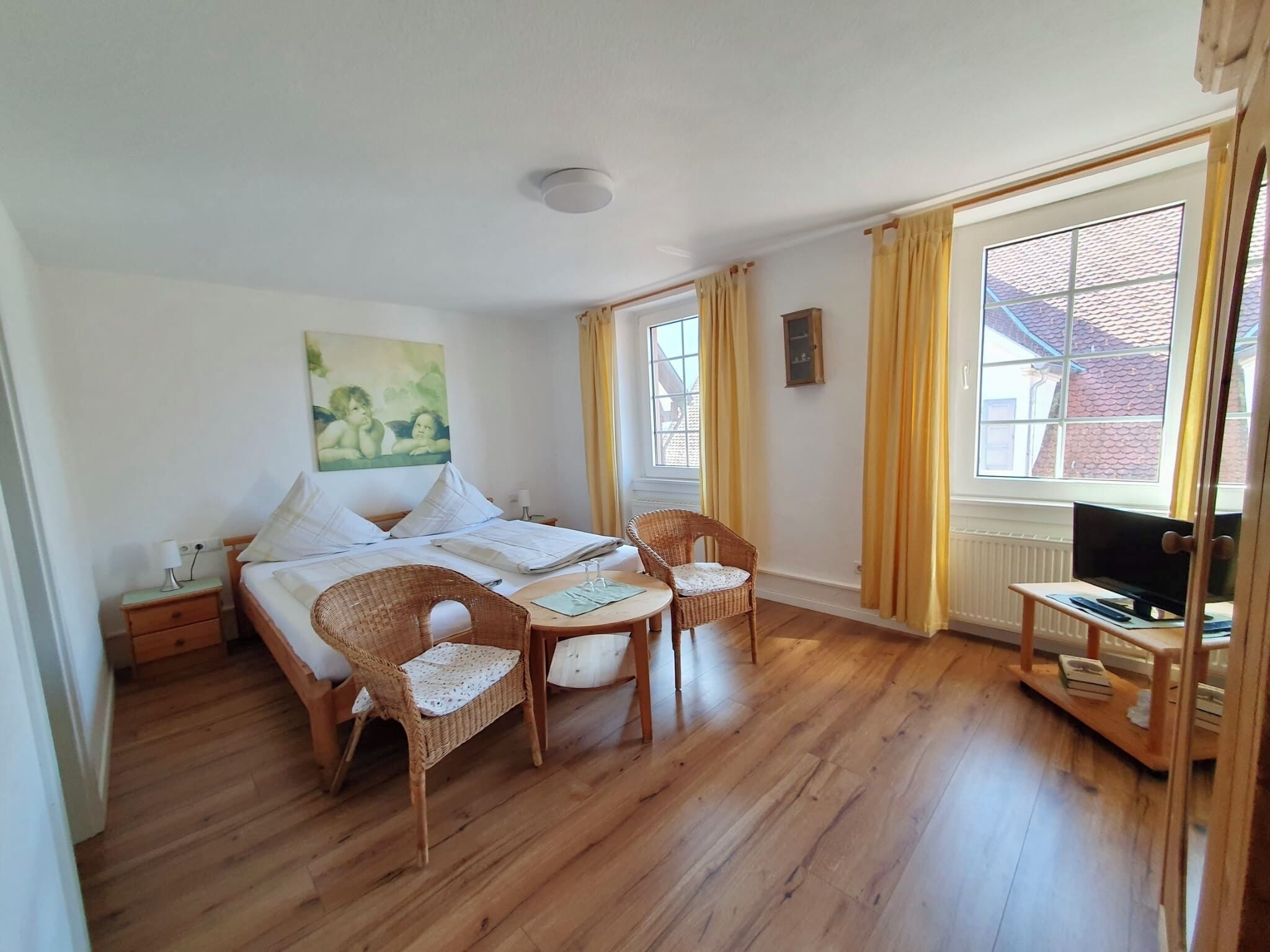 A bedroom with a bed, two wicker chairs, a small table, a TV, and a window with curtains.