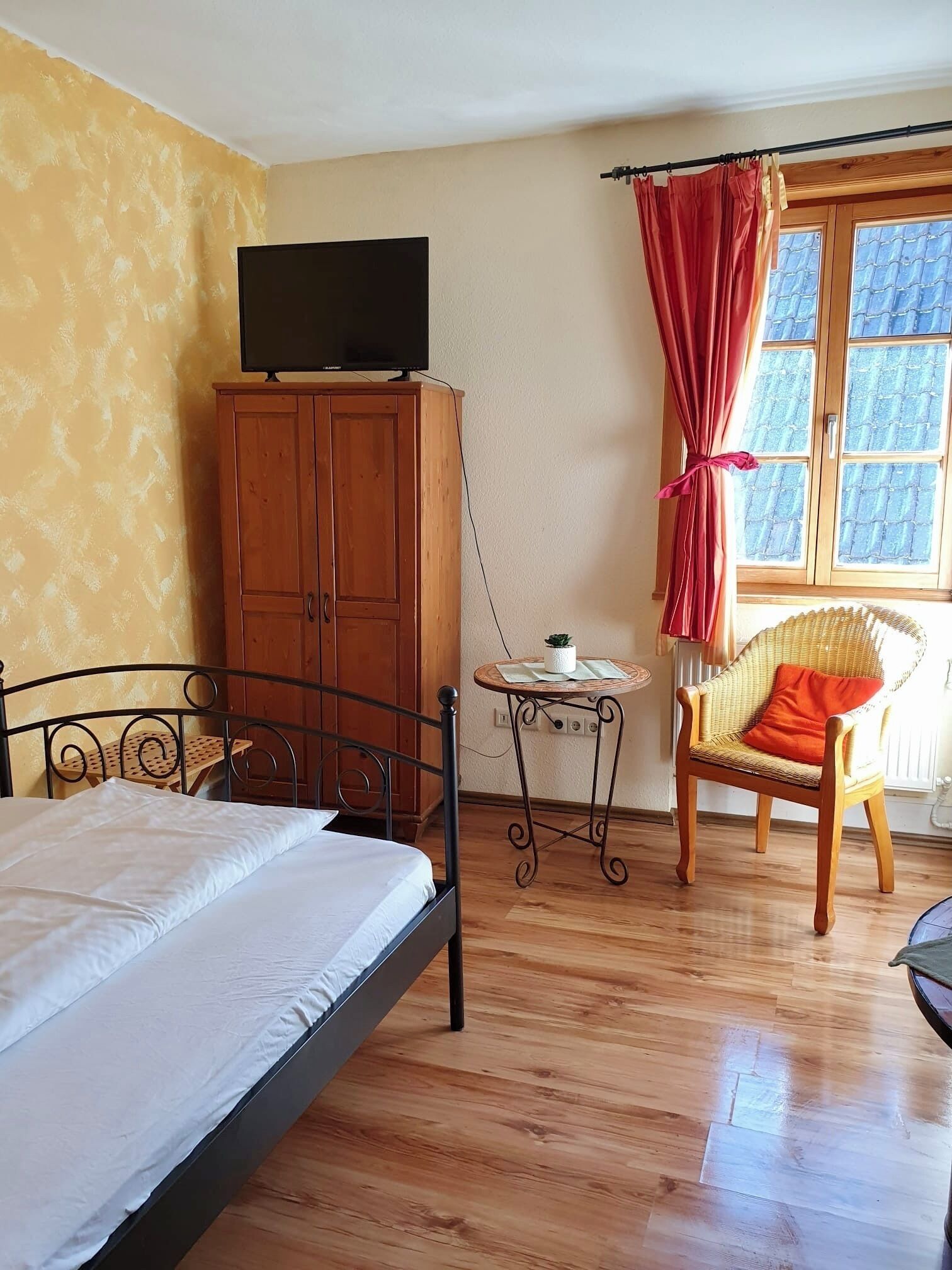 A bedroom with a bed, a wooden wardrobe, a television on top, a small table with a plant, a wicker chair, and a window with curtains.