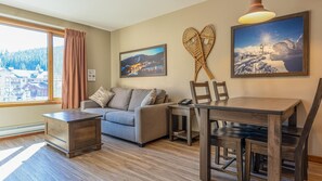 A cozy room with a sofa, a wooden dining table with chairs, a small side table, and a large window with curtains.