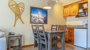 A dining area with a wooden table and chairs, a painting of a snowy landscape, and a kitchen with wooden cabinets and appliances.