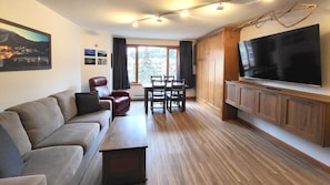 A modern living room with a sofa, a dining table with chairs, a flat-screen TV mounted on the wall, and a large window with curtains.