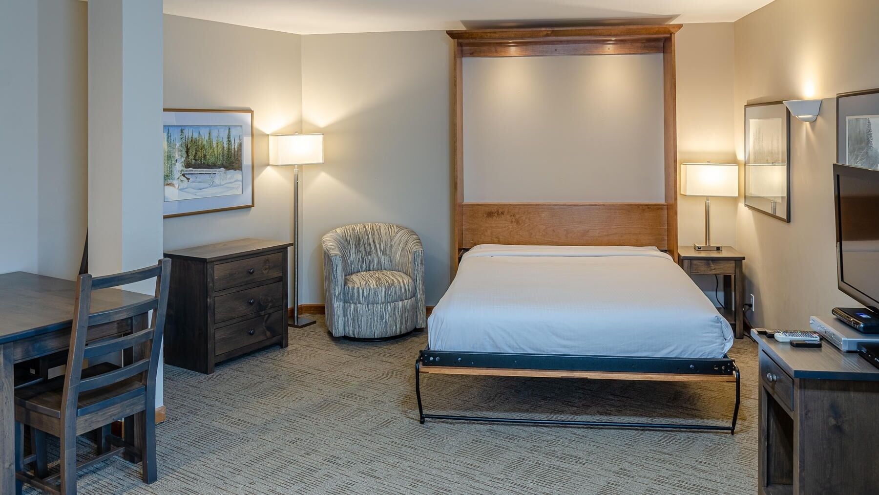 Quarto de hotel com cama, escrivaninha, cadeira, cômoda e televisão.