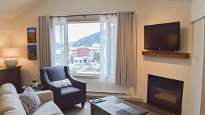 A living room with a sofa, armchair, TV, fireplace, and a large window with curtains.