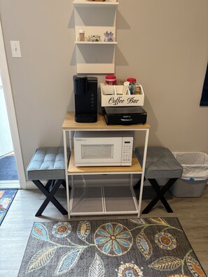 Una kitchenette con cafetera, microondas y una caja de almacenamiento con la etiqueta 'Coffee Bar' sobre una mesa de madera.