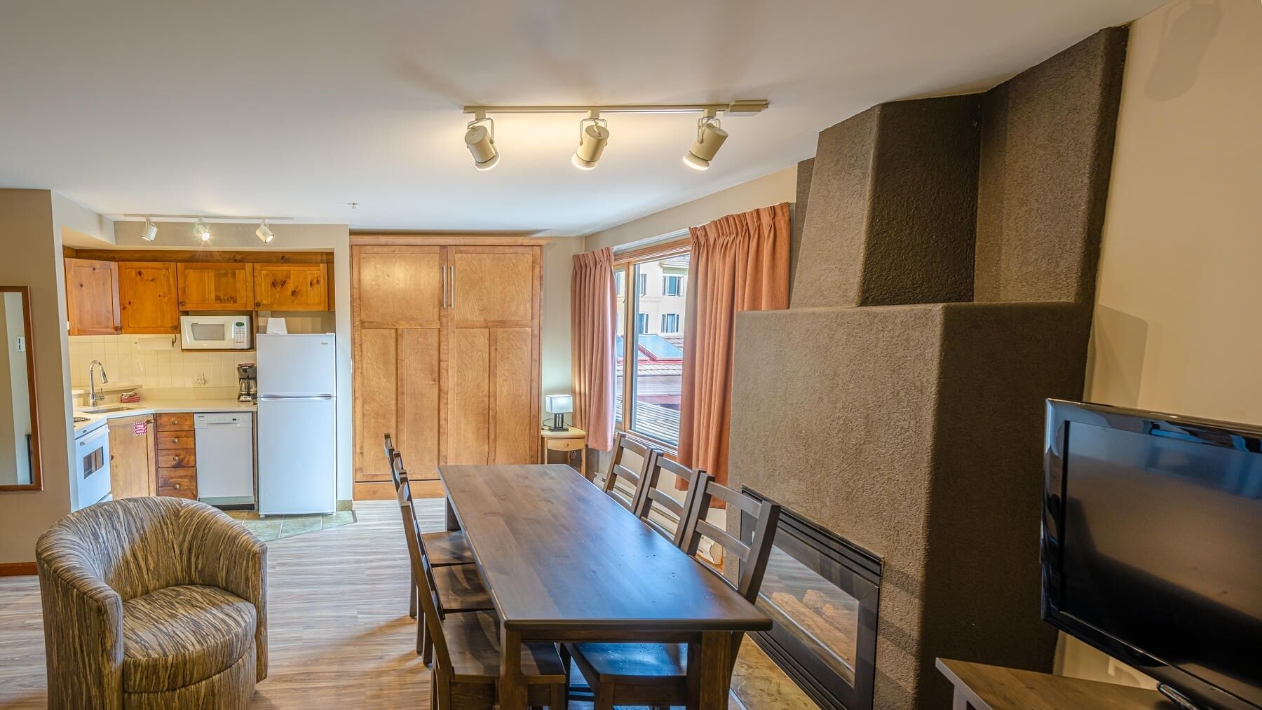 A compact kitchen with wooden cabinets, a microwave, and a refrigerator. There is a dining area with a table and chairs, and a television on a stand.