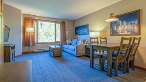 Quarto de hotel com sofá, mesa de jantar e cadeiras, televisão e um quadro na parede.