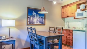 A dining area with a wooden table and chairs, a painting of a snowy landscape, a lamp, and a kitchen with wooden cabinets and a microwave.