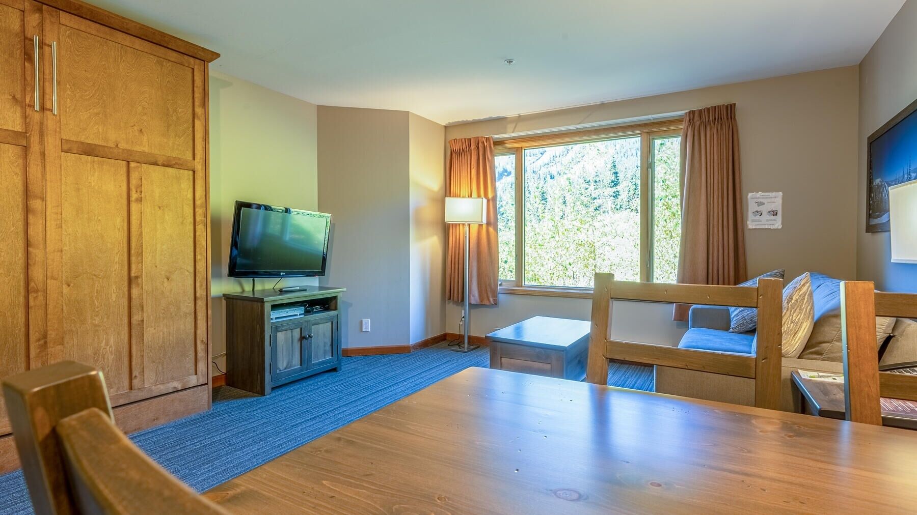 A room with a wooden table, a television on a cabinet, a window with curtains, and a bed with a headboard.