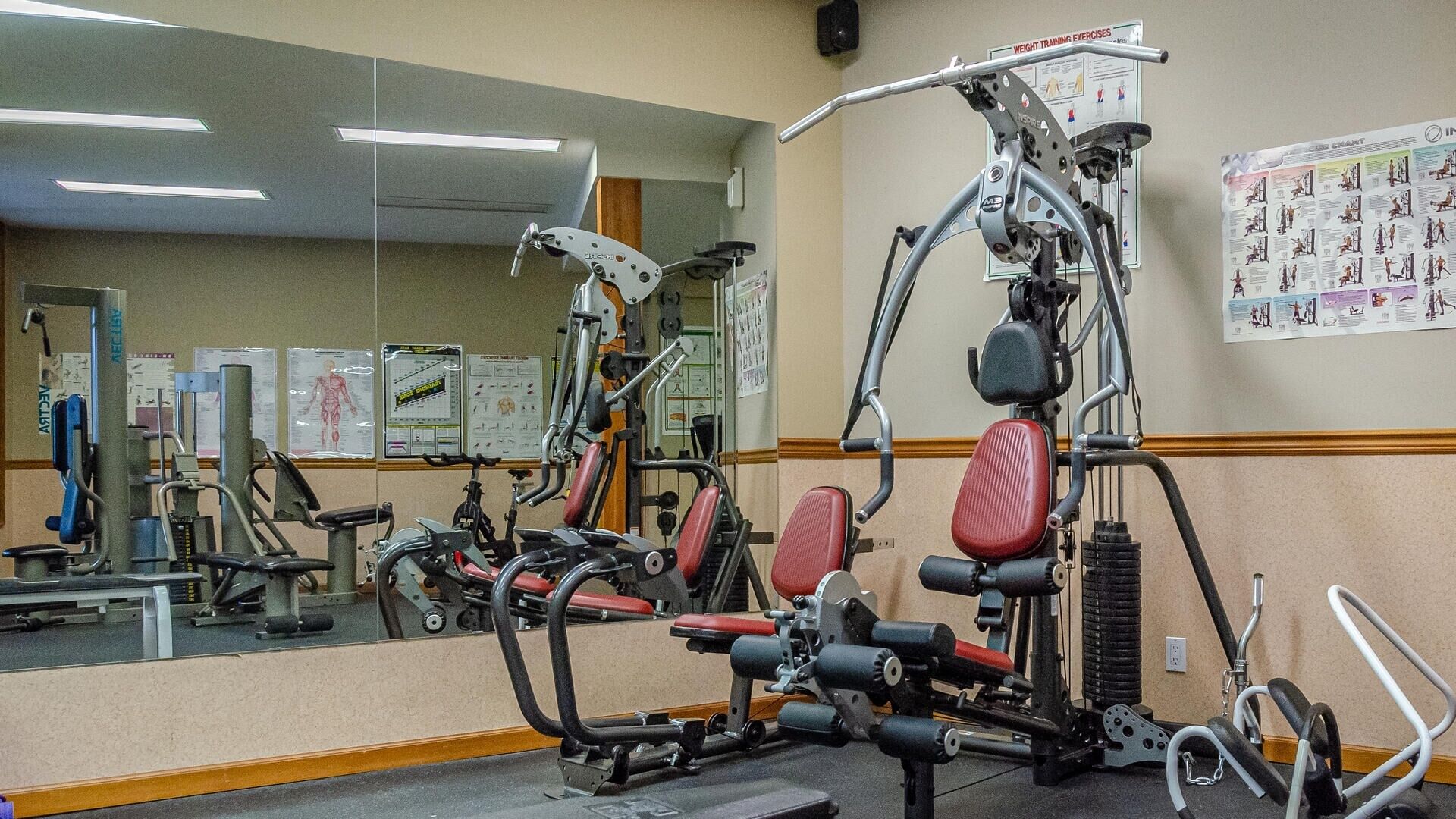 Un gimnasio con diversas máquinas de ejercicio, un espejo grande y carteles instructivos en la pared.