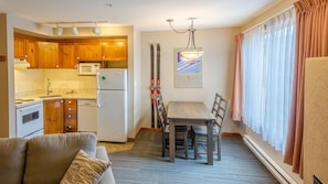 A compact kitchen with wooden cabinets, a microwave, and a refrigerator. There is a dining area with a table and chairs, and a couch in the living room. Skiing equipment is visible against the wall. - Cahilty #332 Hotel Room (Sun Peaks)