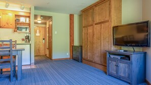 A room with a wooden wardrobe, a TV on a stand, a dining table with chairs, and a kitchen area with appliances. - Cahilty #303 Hotel Room (Sun Peaks)