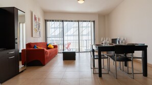 A modern living room with a red sofa, a dining table with chairs, a black cabinet, and a large window with blinds. - The King at Roulette Modern Apartment Paceville (St. Julian's)
