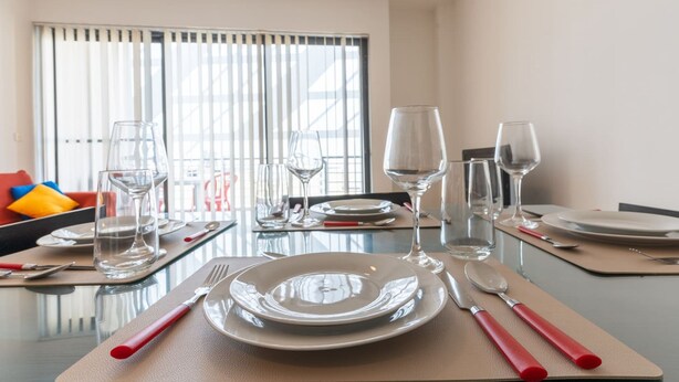 A dining table set with plates, cutlery, and wine glasses, with a sofa and colorful cushions in the background. - The King at Roulette Modern Apartment Paceville (St. Julian's)