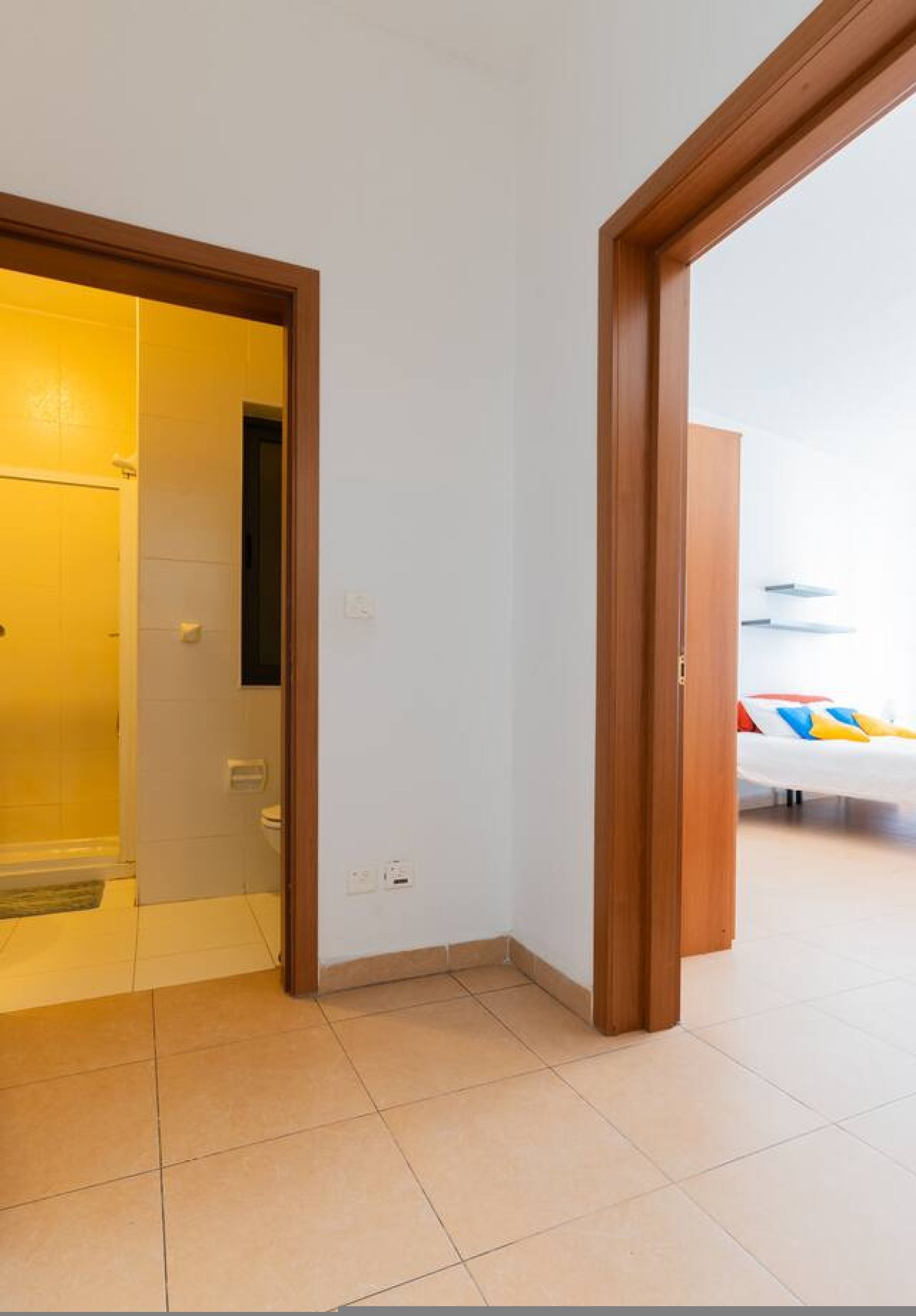 Habitació amb una porta de fusta que dona a un bany, una paret blanca i un llit amb almoades de colors.