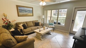 A living room with a beige sofa, a coffee table, and a chair. There is a painting on the wall and a ceiling fan. - Nothing Between You & The Beach 2 Bedroom Condo (South Padre Island)