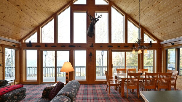 A lodge interior with a high ceiling, large windows, a dining area, and a mounted deer head. - Loon Song On Lost Land Lake 4 Bedroom Home (Hayward)