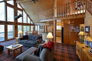 A cabin interior with a loft bedroom, a dining area, and a living room with plaid flooring.