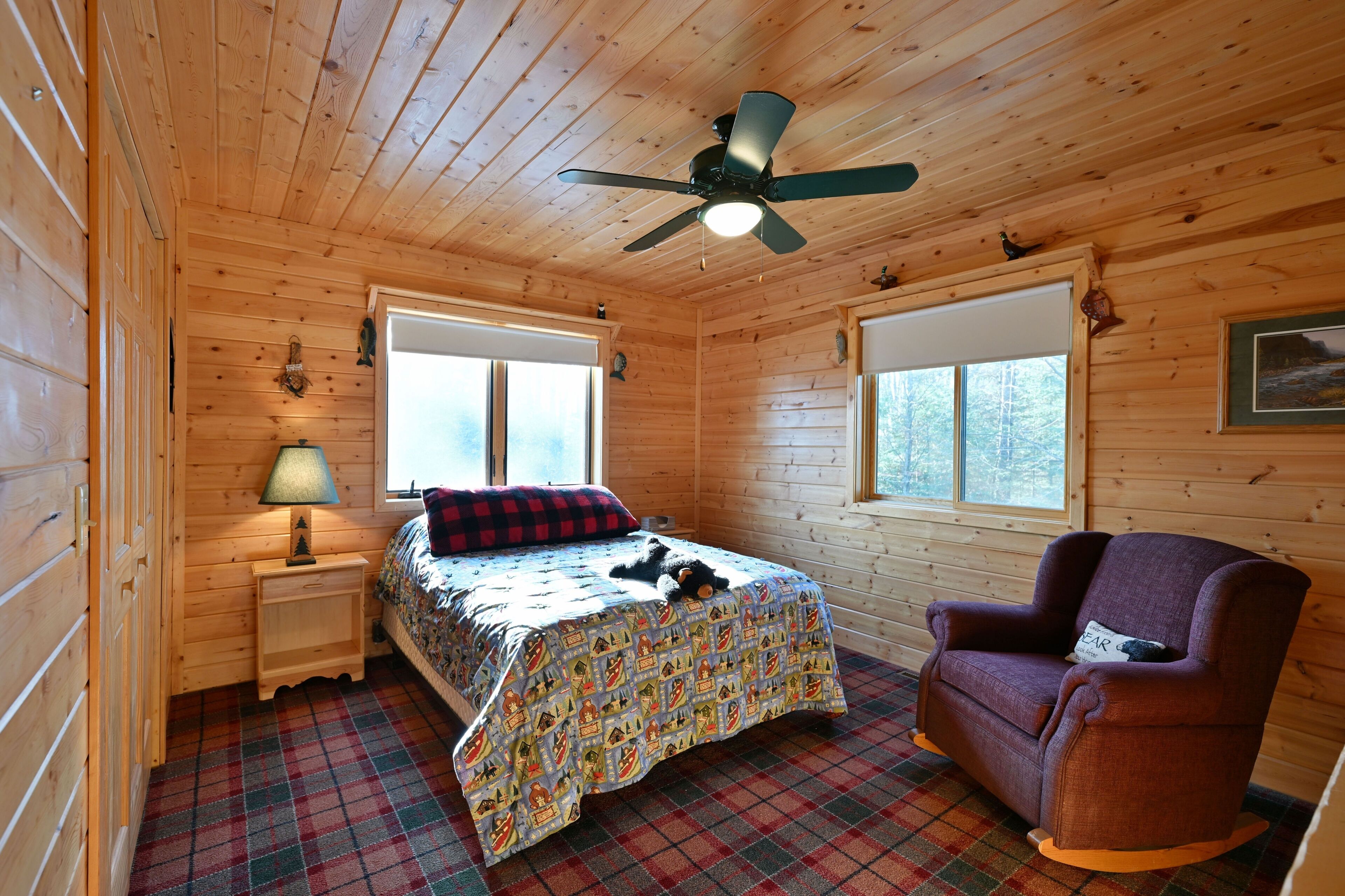 A cozy cabin room with a bed, a chair, a ceiling fan, and two windows.