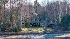 A lakeside cabin with a deck and a covered porch, surrounded by trees. - Loon Song On Lost Land Lake 4 Bedroom Home (Hayward)