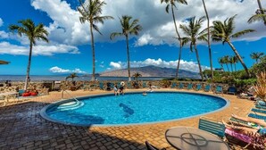 A swimming pool with lounge chairs and palm trees, surrounded by a stone wall and a view of the ocean. - Menehune Shores, #522 2 Bedroom Condo (Kihei)