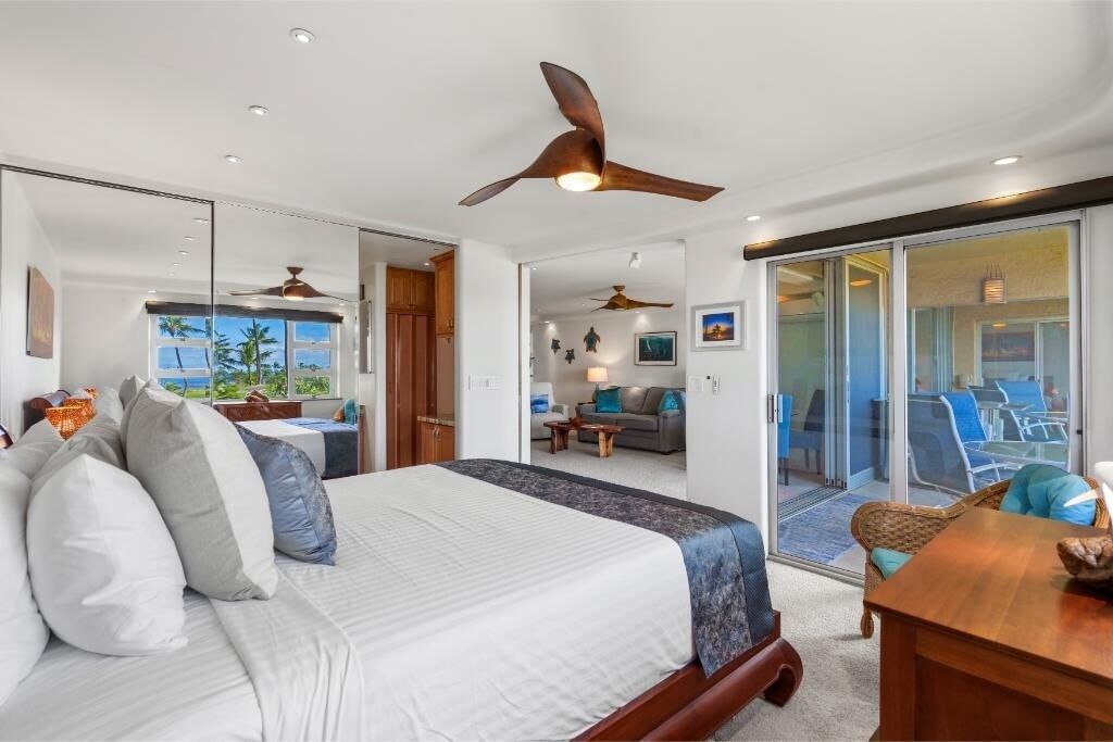 A hotel room with a bed, ceiling fans, a balcony with chairs, and a view of the outdoors.