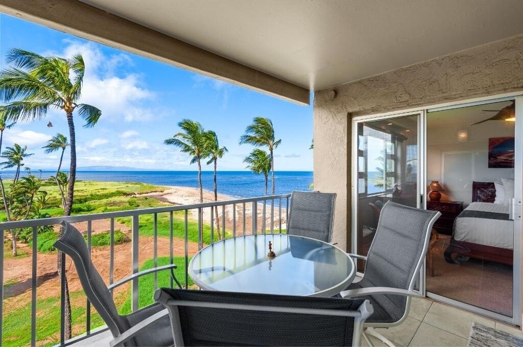 A balcony with a glass table and chairs, overlooking a beach with palm trees and the ocean.