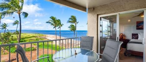 A balcony with a glass table and chairs, overlooking a beach with palm trees and the ocean.