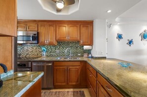 A kitchen with wooden cabinets, a stainless steel microwave, a sink, and a backsplash with a mosaic tile pattern. - Menehune Shores, #522 2 Bedroom Condo (Kihei)