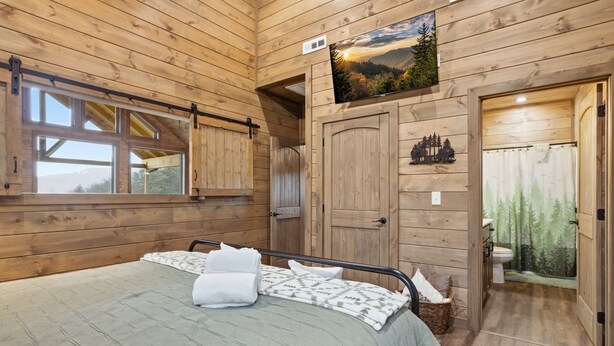 Ein Zimmer in einer Holzhütte mit einem Bett, einem Fenster, einer Tür und einem an der Wand befestigten Fernseher.